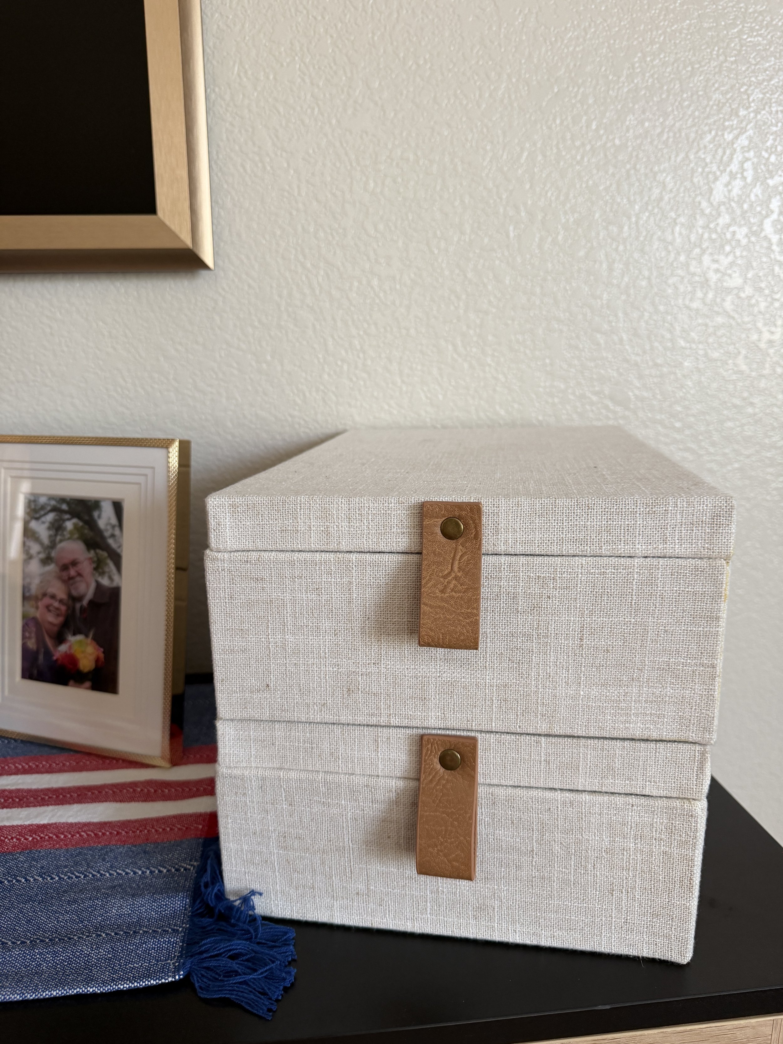 Two stacked linen boxes with leather tab closures holding printed family photos, displayed beside a framed portrait