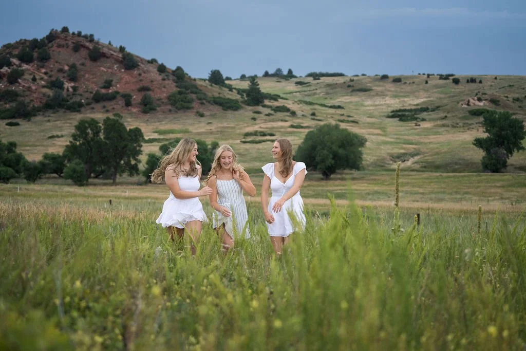Three high school senior girls laughing in a Colorado meadow with red rock hills — Castle Rock senior photographer Jennie Bennett Photography