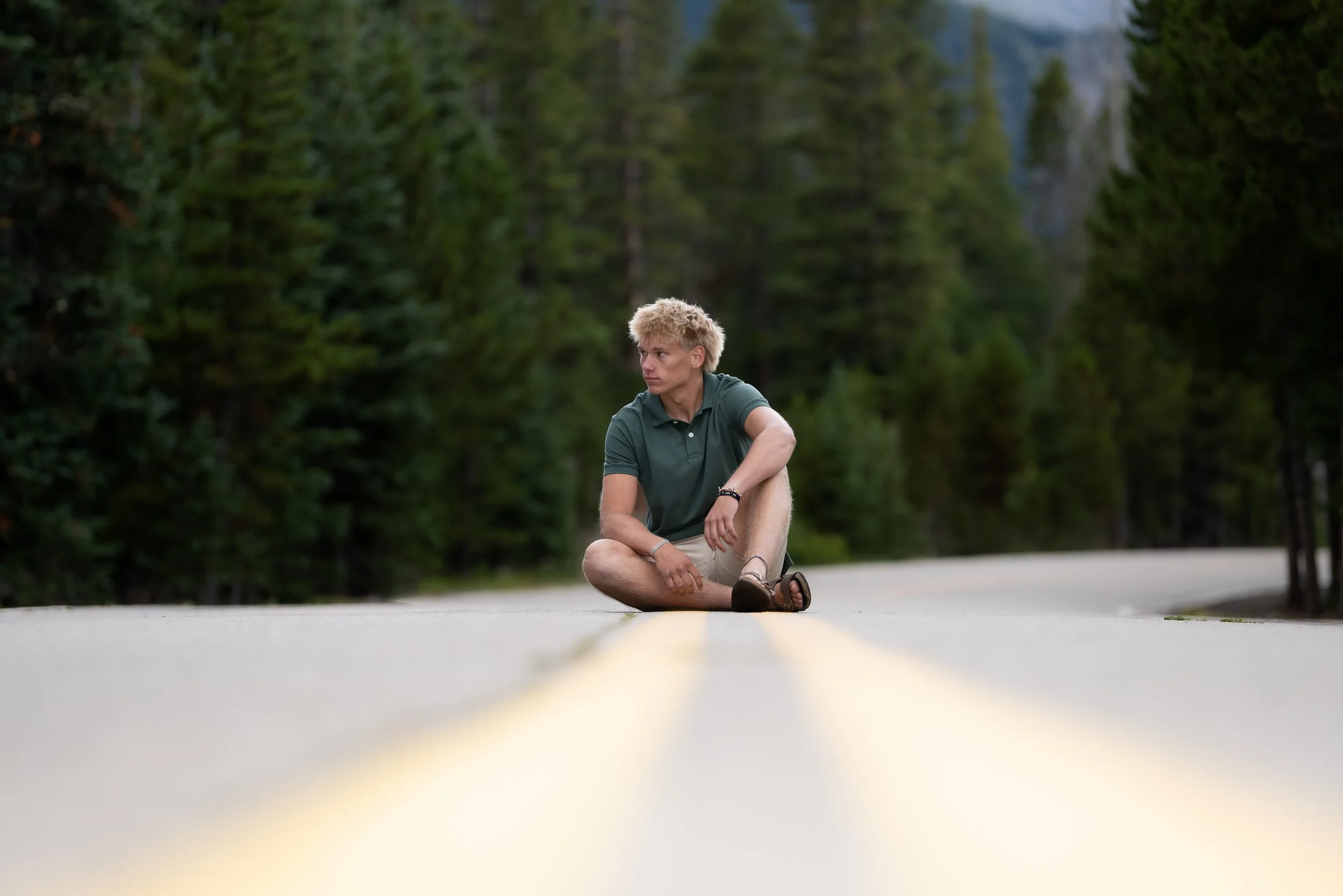 High school senior guy sitting on a quiet mountain road surrounded by pine trees, Colorado senior portrait session