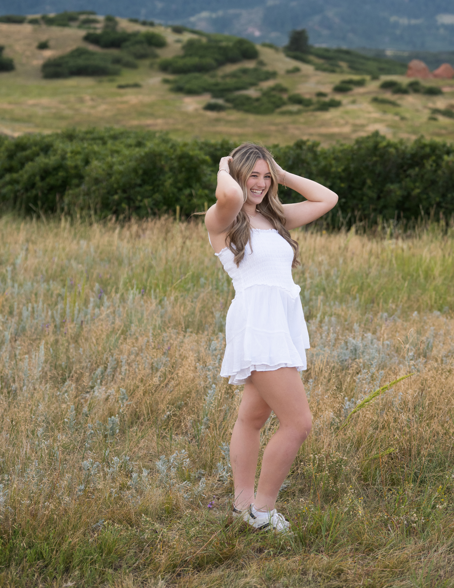High school senior girl in white dress smiling in golden hour meadow with Colorado mountains — Castle Rock senior photographer Jennie Bennett Photography
