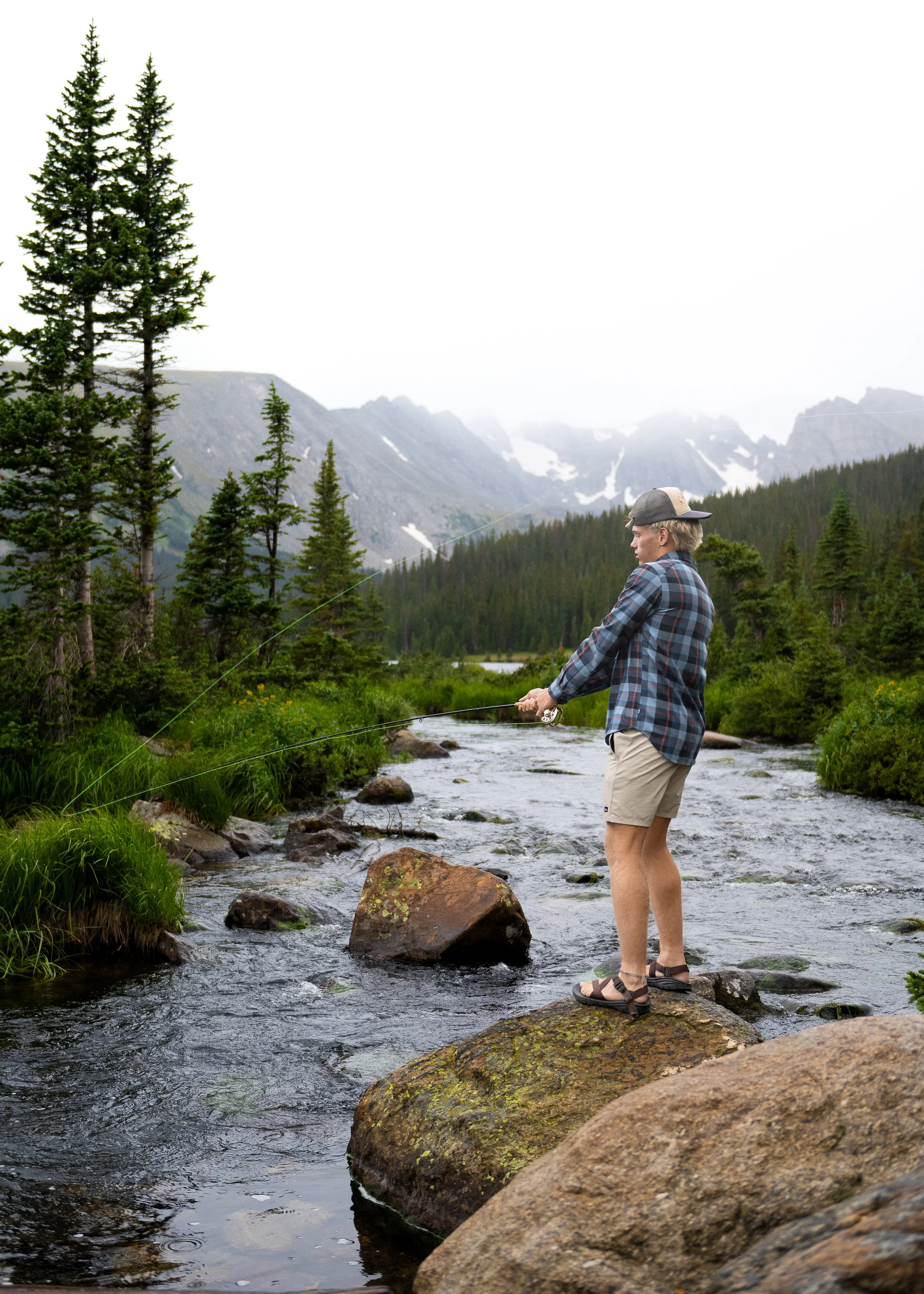 High school senior guy fly fishing from a boulder in a Colorado mountain stream with snowy peaks behind him, senior pictures by Jennie Bennett Photography