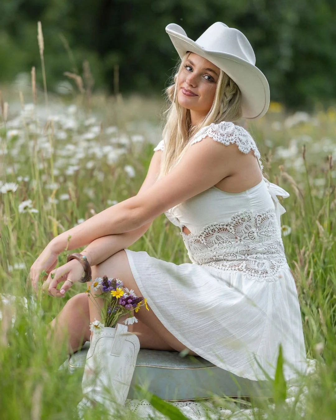 High school senior girl in a white lace dress and cowboy hat sitting in a field of wildflowers holding a bouquet, Colorado senior photos by Castle Rock photographer Jennie Bennett Photography