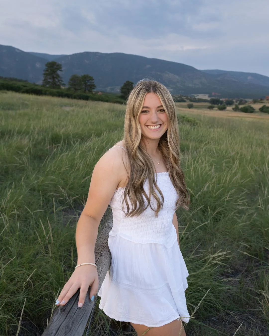 High school senior girl smiling in a white romper leaning on a wooden fence post with a green meadow and Colorado mountains in the background, photographed by Jennie Bennett Photography