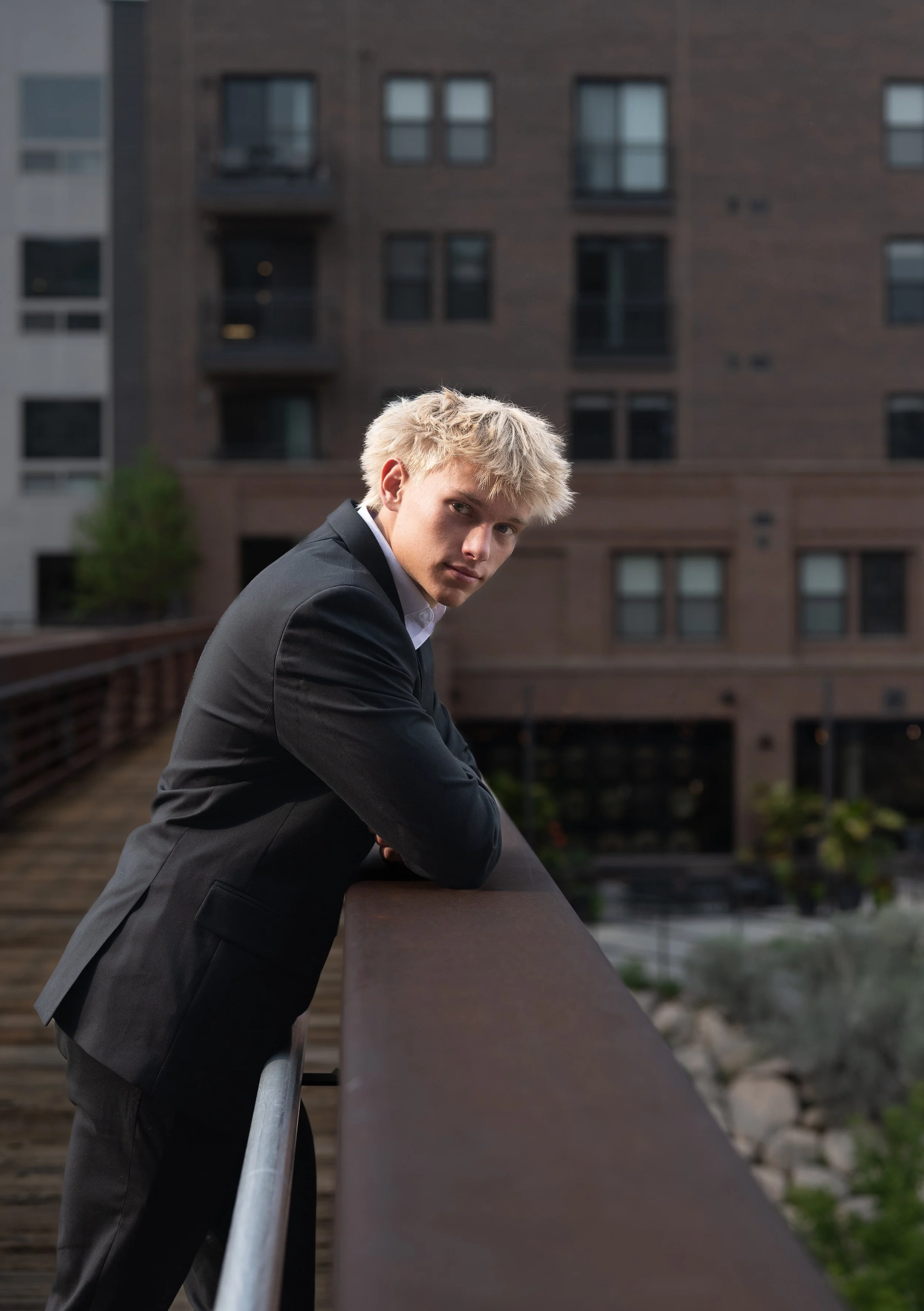 High school senior guy in a dark suit leaning on a railing with city buildings behind him, urban senior photos in Denver by Castle Rock photographer Jennie Bennett Photography