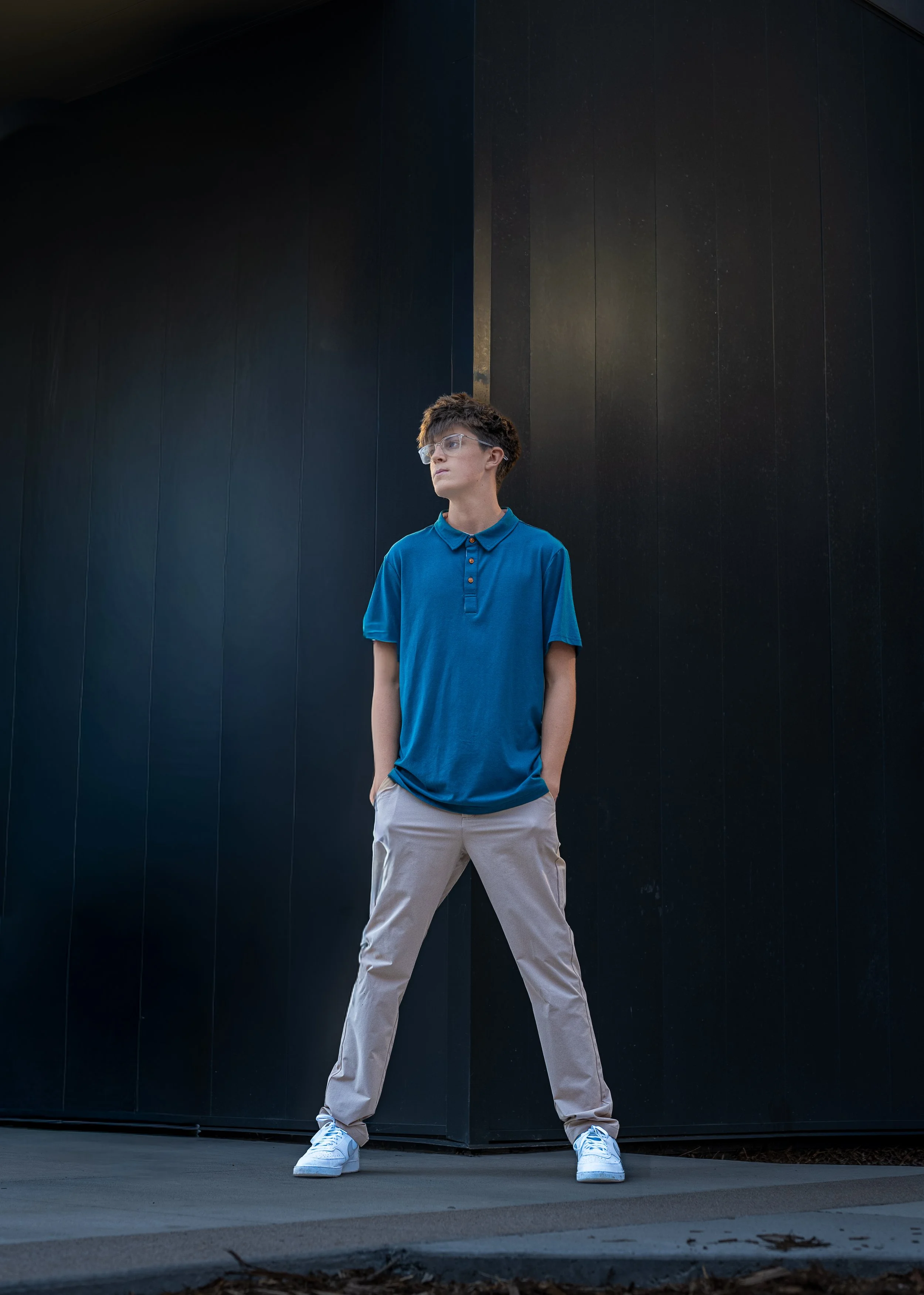High school senior guy in a blue polo standing confidently against a dark black wall, urban Colorado senior portraits by Jennie Bennett Photography