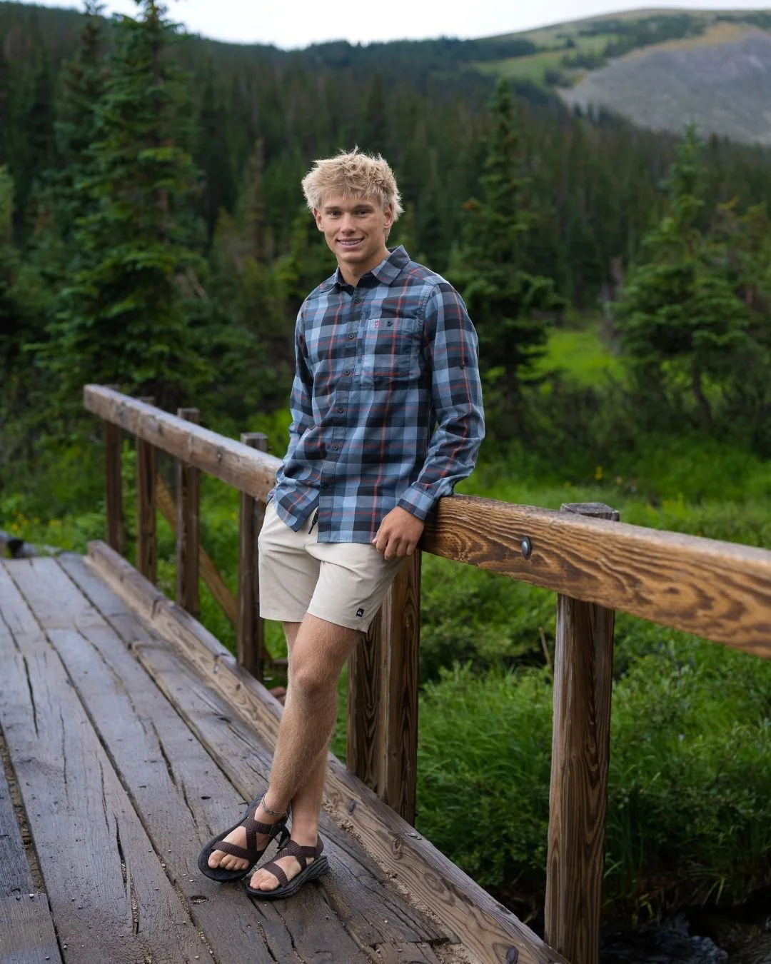 High school senior guy in a plaid shirt leaning on a wooden footbridge in a green mountain forest, Colorado senior photos by Castle Rock photographer Jennie Bennett Photography