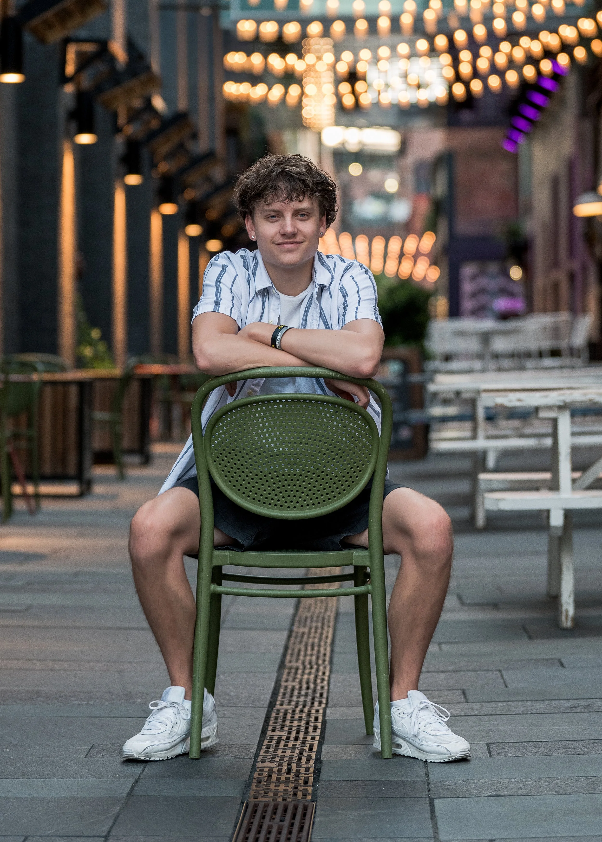 High school senior guy sitting backwards on a green chair under glowing string lights on a city patio, Denver senior pictures by Jennie Bennett Photography