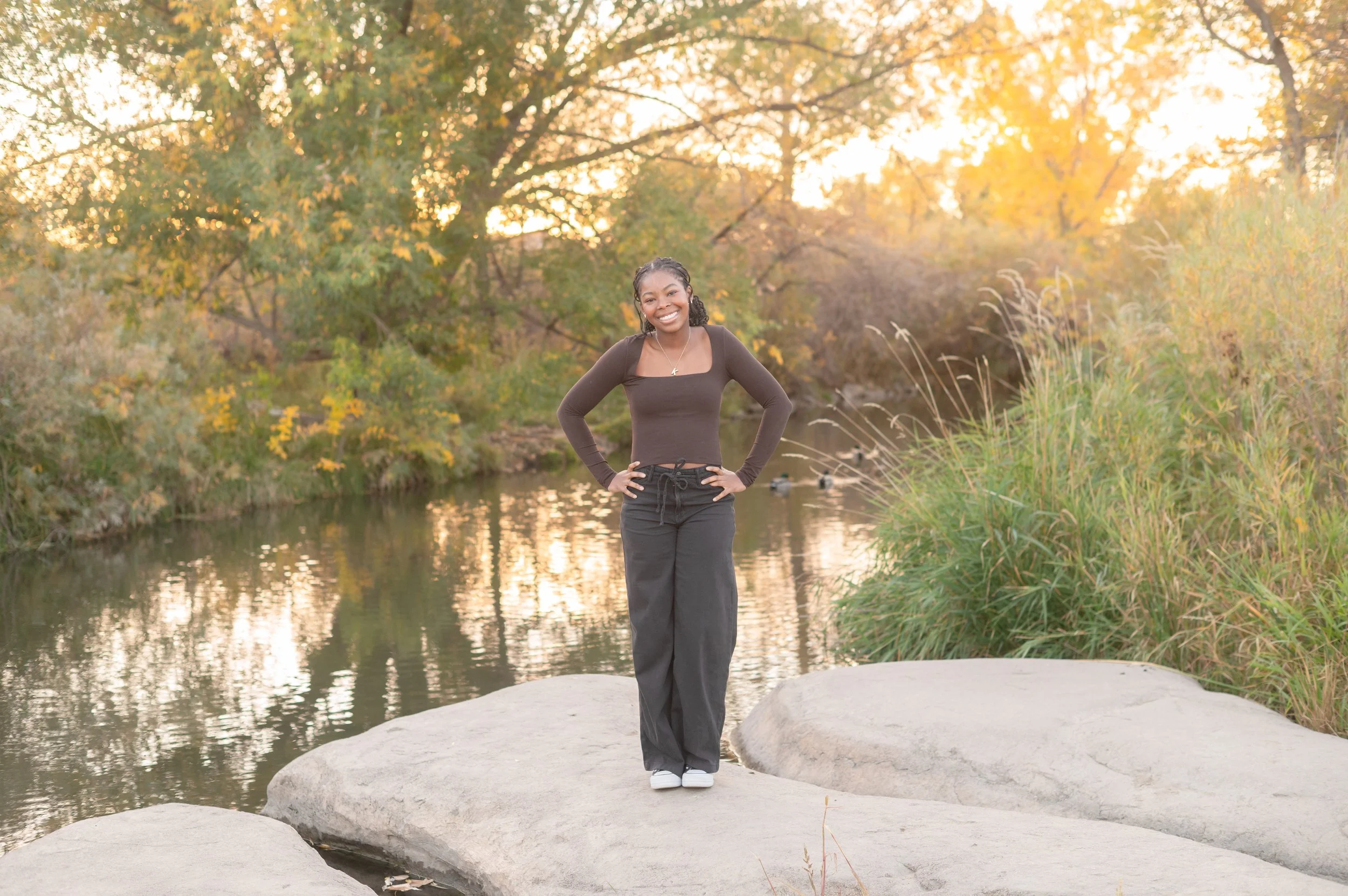 High school senior girl smiling at golden hour standing on  rocks by a creek with fall foliage — Denver senior portrait  photographer Jennie Bennett Photography