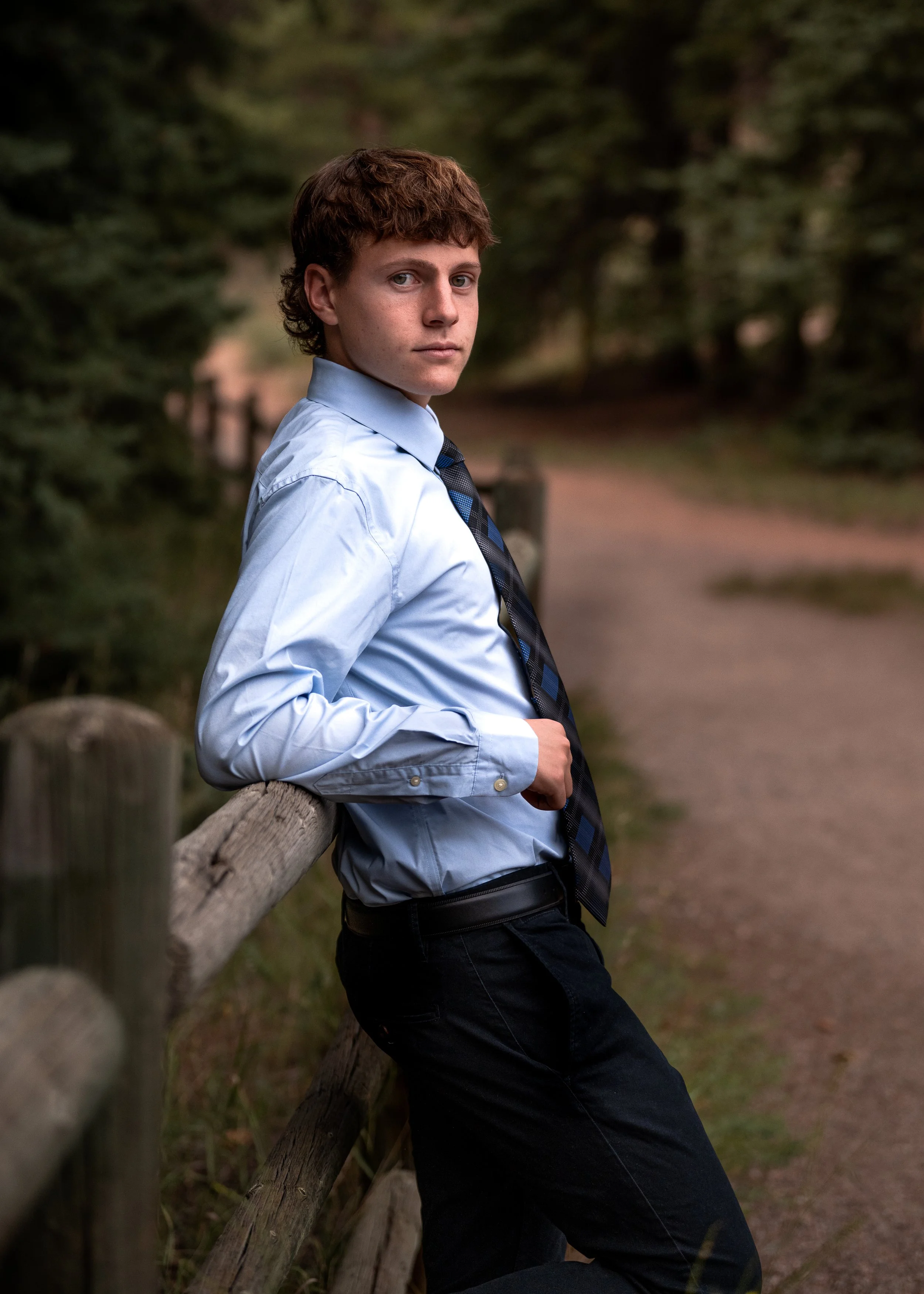 High school senior guy in a light blue shirt and tie leaning on a wooden fence along a pine forest trail, Colorado senior pictures by Jennie Bennett Photography