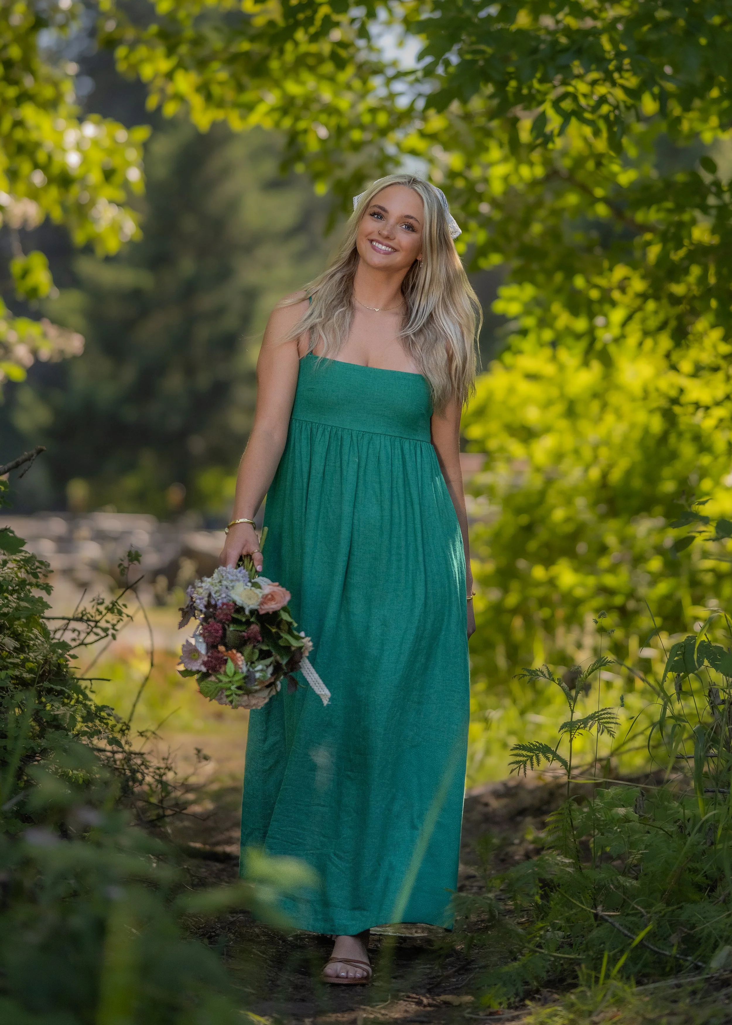 Denver senior girl in a green dress walking through greenery and holding a bouquet during her senior portrait session, smiling at the camera.