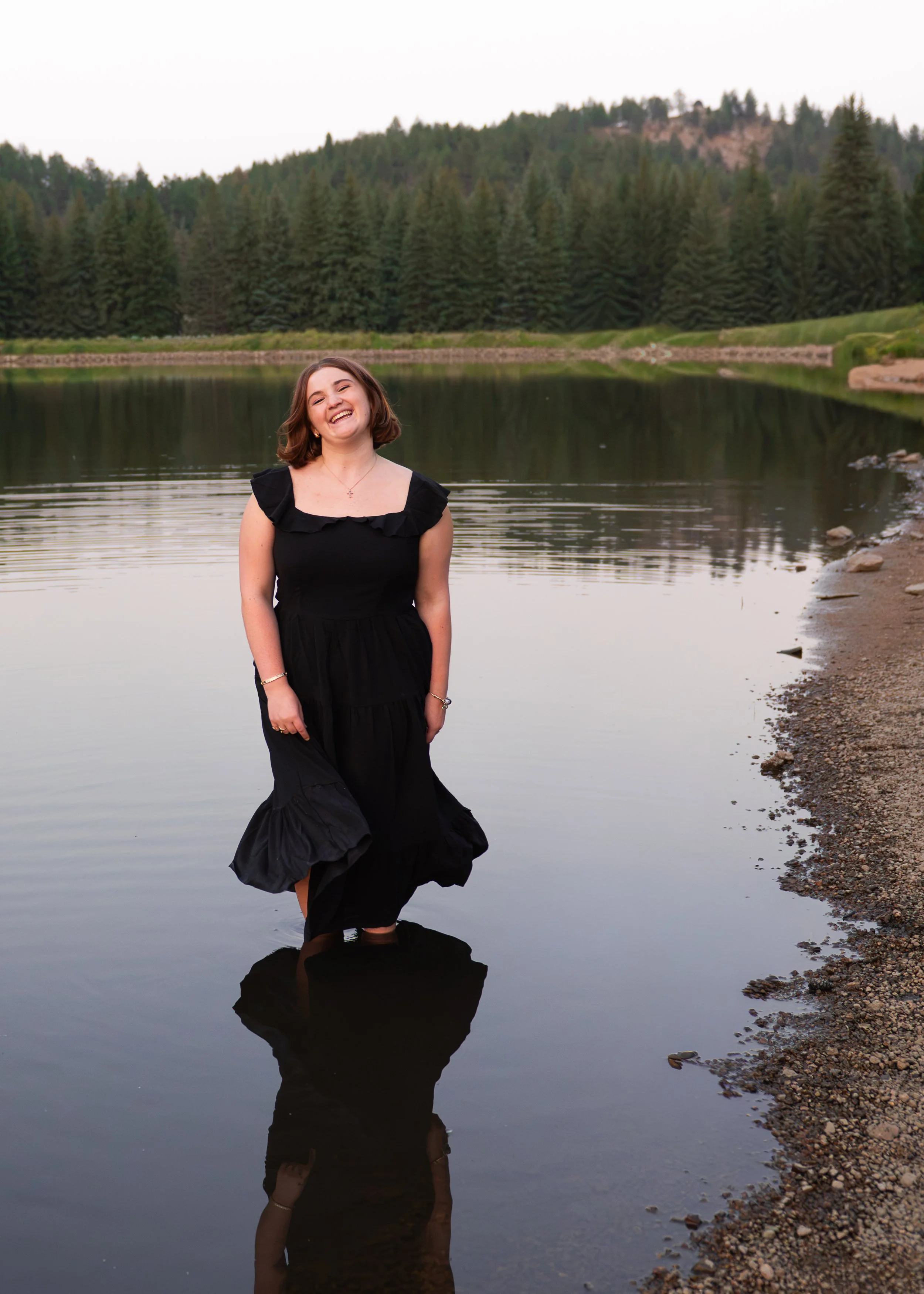 High school senior girl in a black dress laughing while standing in a mountain lake surrounded by pines, Colorado senior portraits by Jennie Bennett Photography