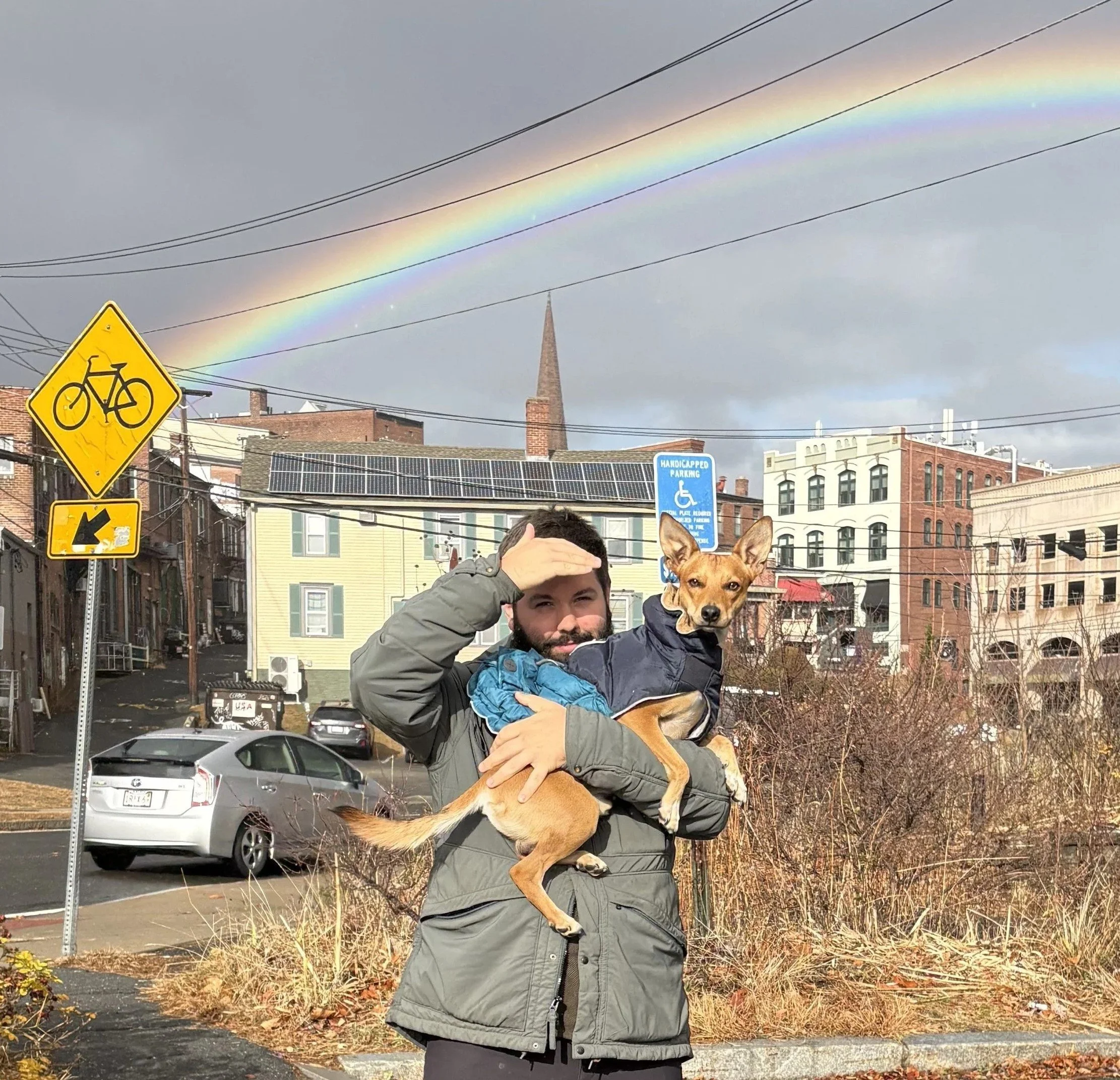 A man stands in an urban landscape under a rainbow while holding a small dog.