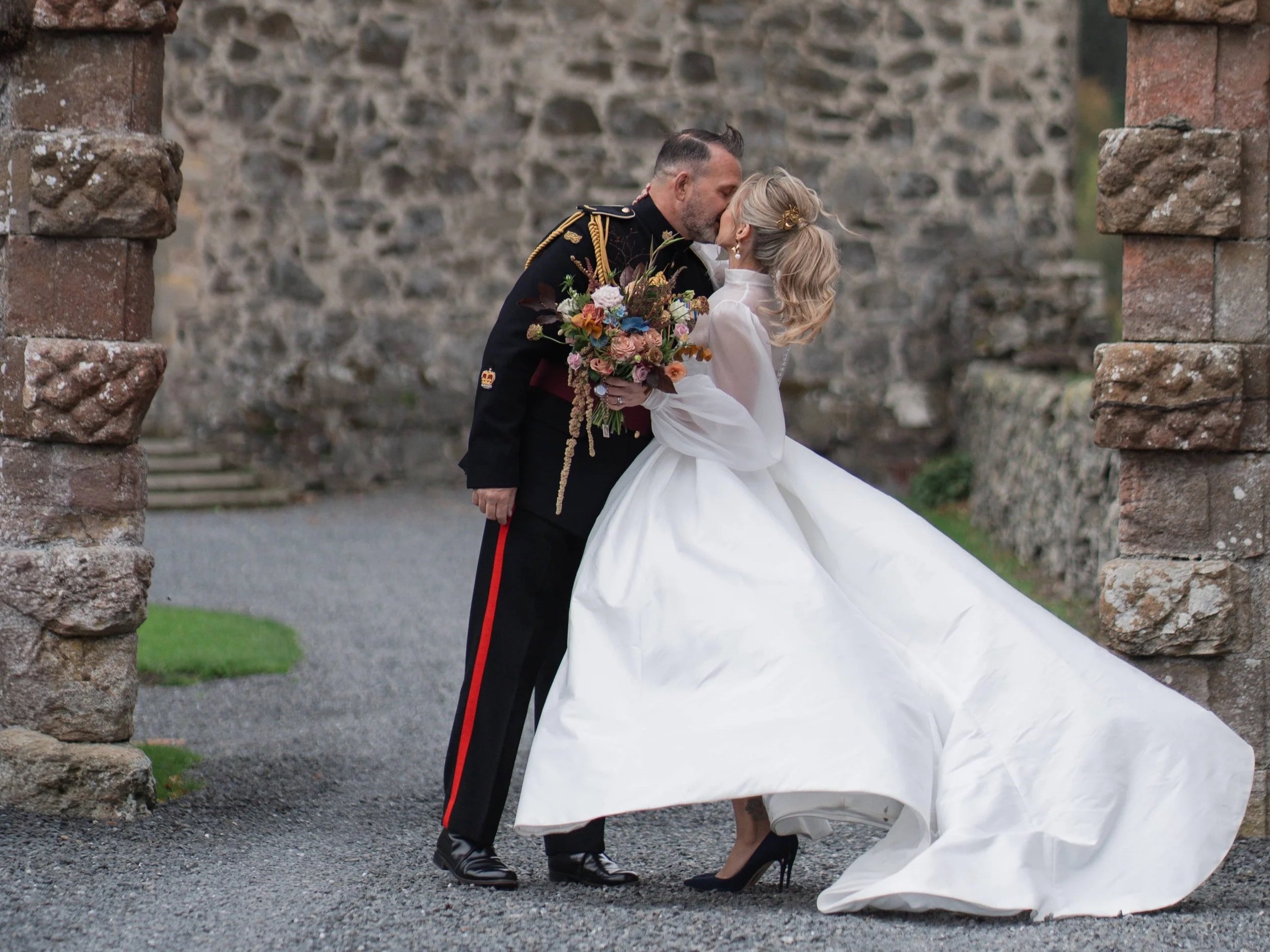 Jaymie and Karl share a kiss outside the castle. Jaymie holds her bouquet and the wind blows the train of her dress.