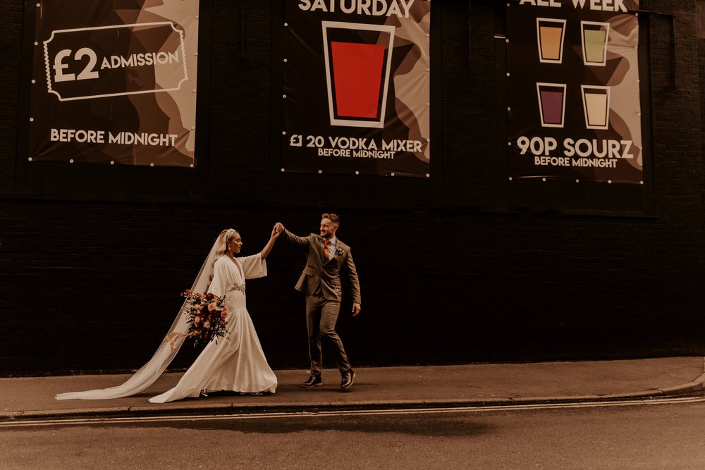 Lucy and her husband Jody dance on the pavement next to the wall of a large black building