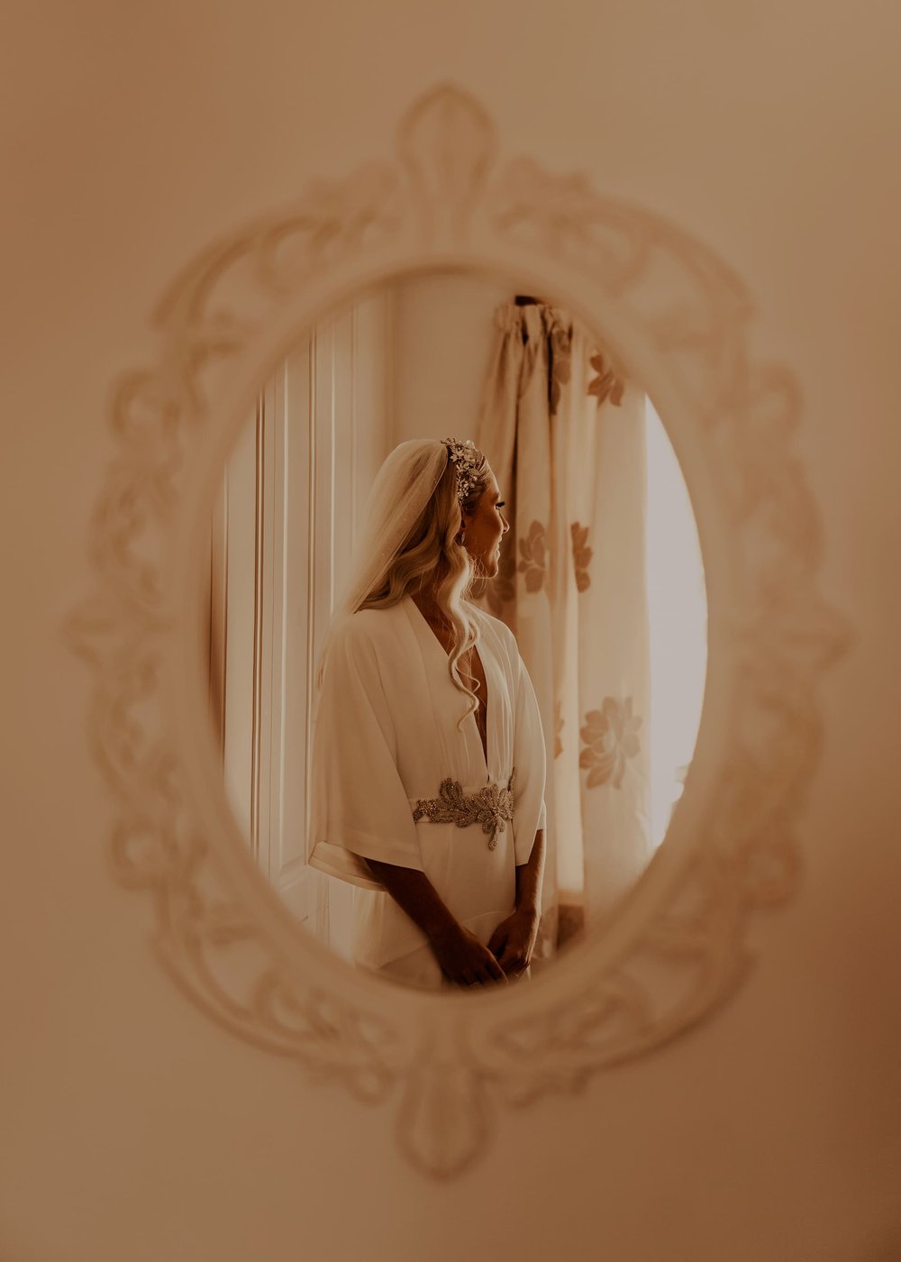An image of a mirror showing Lucy looking out of a window, in her wedding dress, headband, belt and veil.