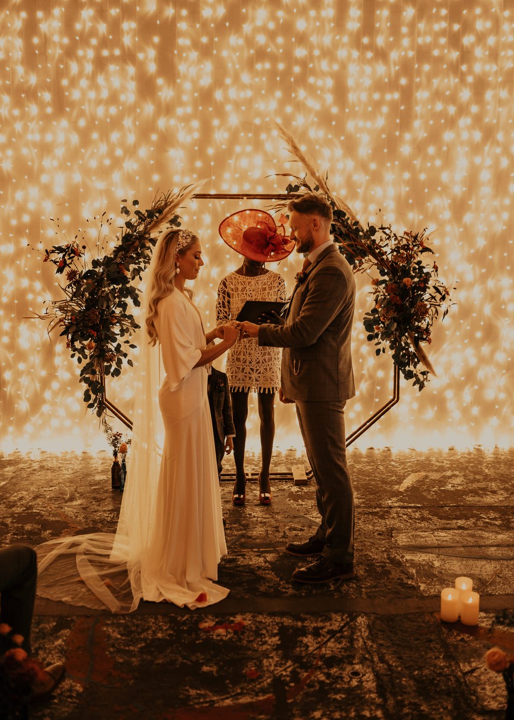 Lucy and Jody stand holding hands during their ceremony, led by a celebrant. The backdrop is a wall full of fairy lights and a foliage installation.