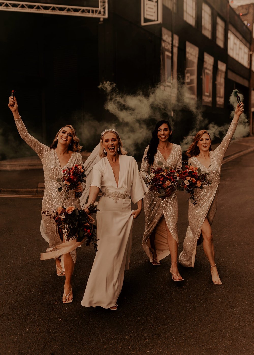 Lucy walks down a road with 3 bridesmaids who are wearing gold sequinned long sleeve dresses. They're carrying their bouquets and waving smoke bombs