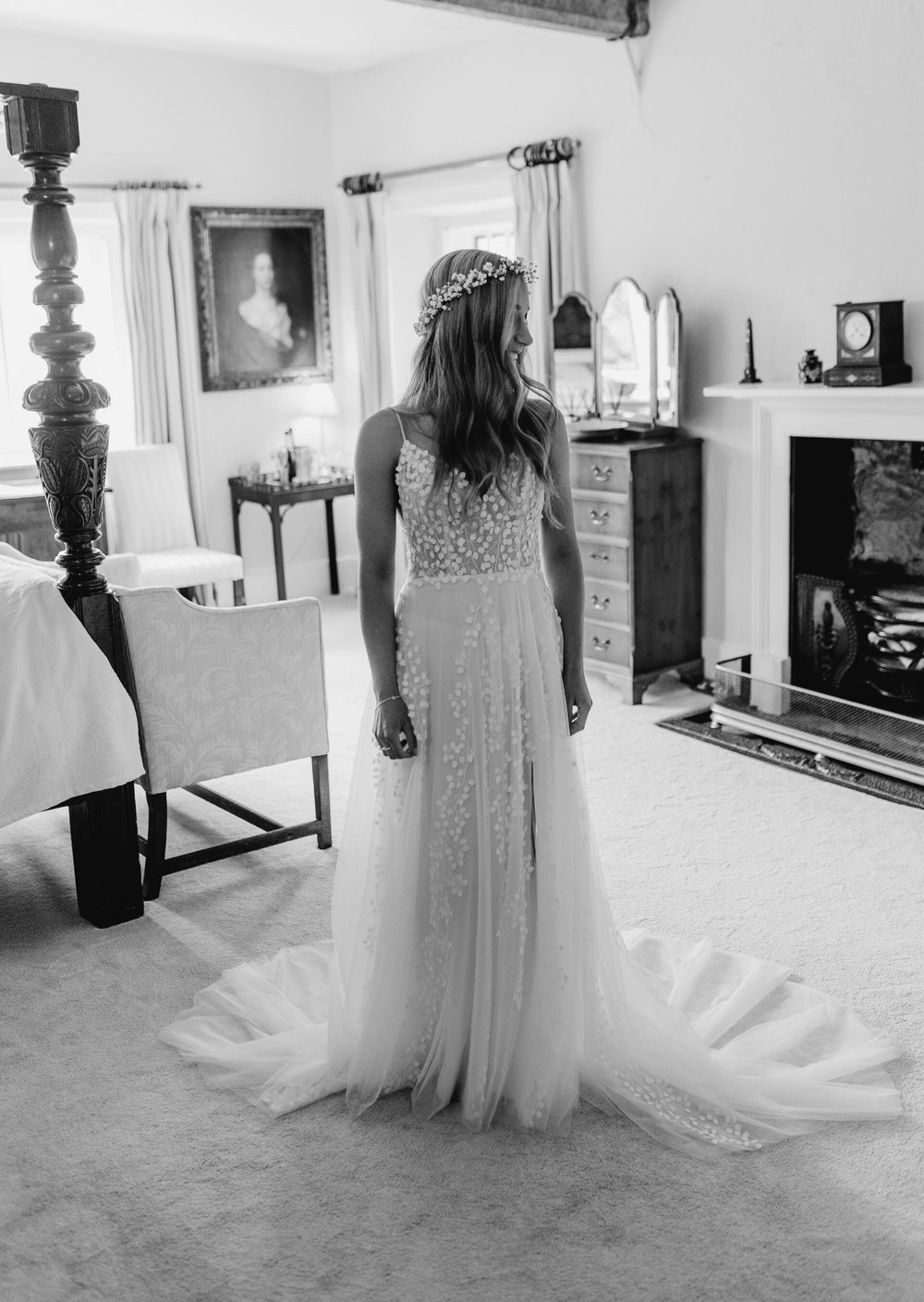 A black and white photo where Alice stands in a room at Eyam hall, looking over her shoulder. She wears her wedding dress, headband and has loosely curled hair left down.