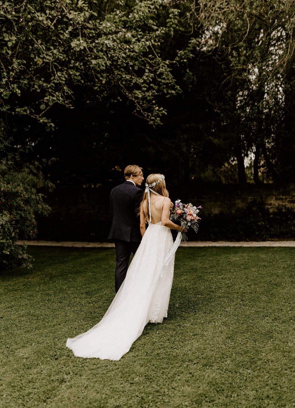 Alice and Conor walk holding hands through Eyam Hall grounds, with her dress train flowing behind her. The dress has a low, open back.