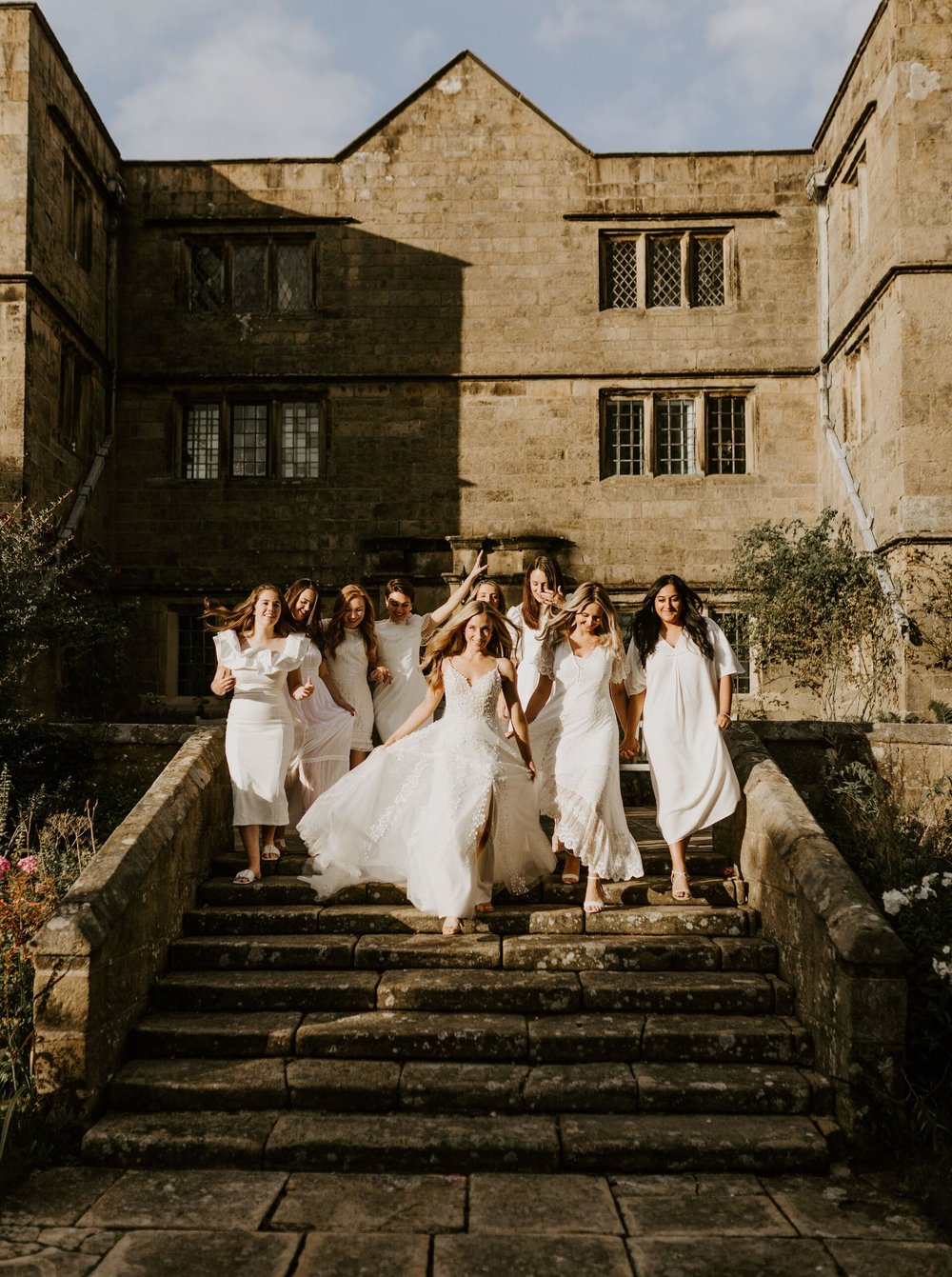  Alice walks down a set of wide stone steps in front of historic Eyam Hall, closely followed by her 8 bridesmaids in white dresses.  