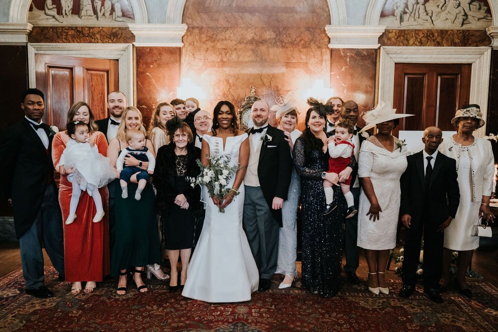  Meisha and Patrick stand in a room of the hall surrounded by friends and family for a large group photo. 