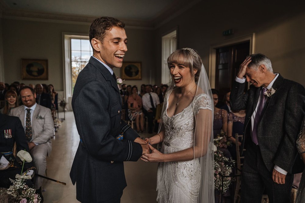  Sam and Emma stand at the top of the aisle for their vows. They are both laughing whilst holding hands facing each other. The ceremony is in a large room with georgian style windows and panelling.  