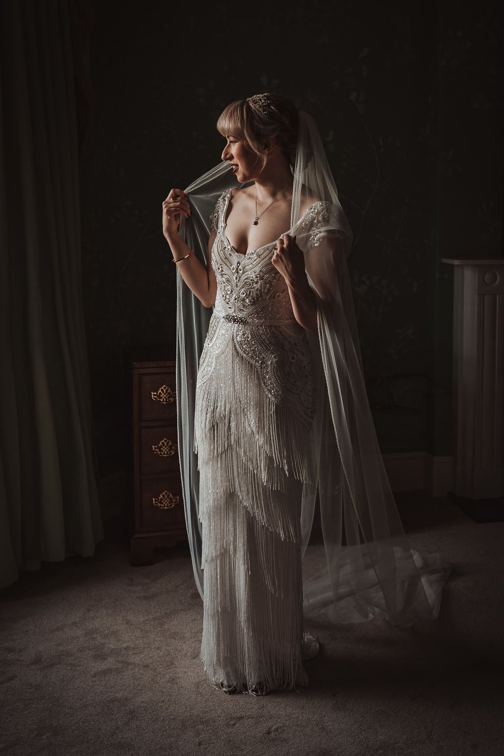  A portrait image of Emma who stands facing the camera with her head turned towards natural light from a window. She holds her veil around her shoulders. You can see the full length of the dress, with detailed ivory beading and tassels from the hip t