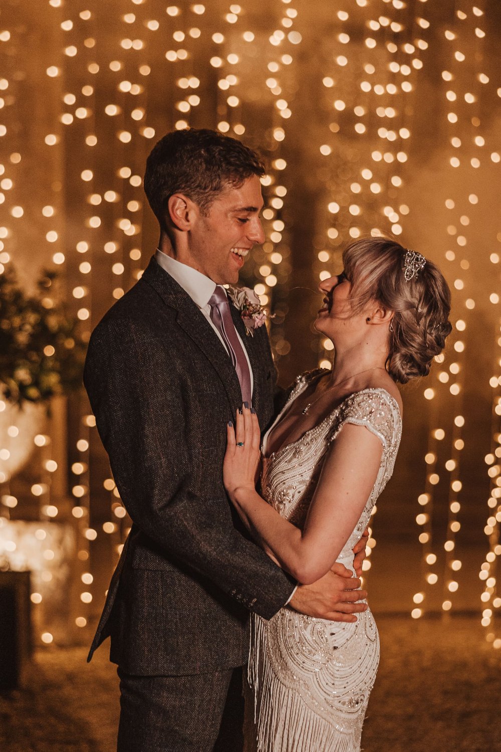  Sam and Emma stand in front of a waterfall of fairy lights. They are looking at each other, embracing and laughing.  