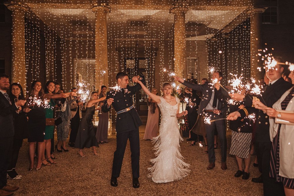  Sam and Emma walk through two lines of their guests, all holding sparklers now it is dark outside. Behind them all is a waterfall of fairy lights hanging on the outside of the building. 