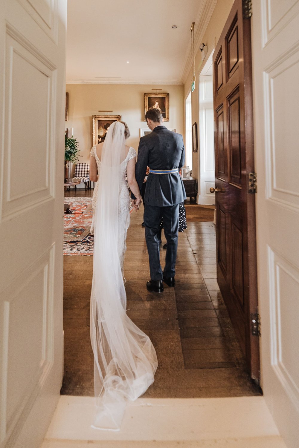  Emma and Sam walk hand in hand through a panelled door into a large drawing room of their venue. They are walking away from the camera and you can see the full floor length of Emma’s veil. 