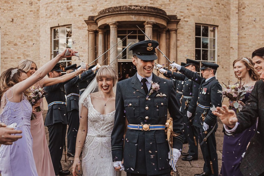  Emma and Sam walk hand in hand towards the camera underneath a military sword arch after the wedding ceremony.  