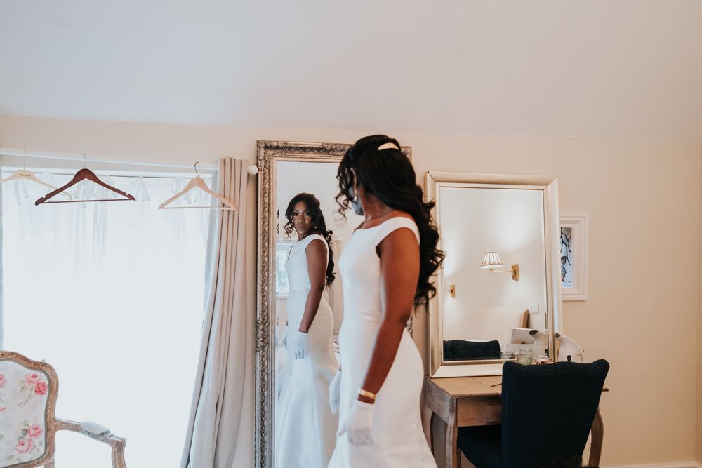  Meisha looks at herself in a full length mirror, wearing her wedding dress with hair and make up done. The dress is a satin fishtail sleeveless dress, with a wide boat neck. 