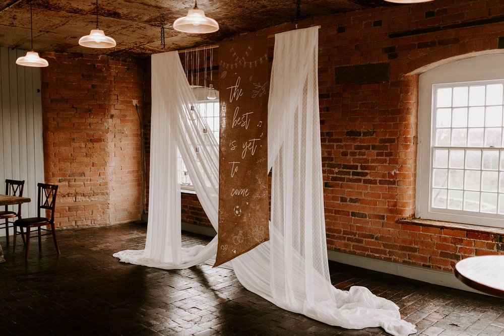  A handprinted sign hangs in the venue, styled in front of some sheer white material that hangs down. The sign reads ‘the best is yet to come’. 