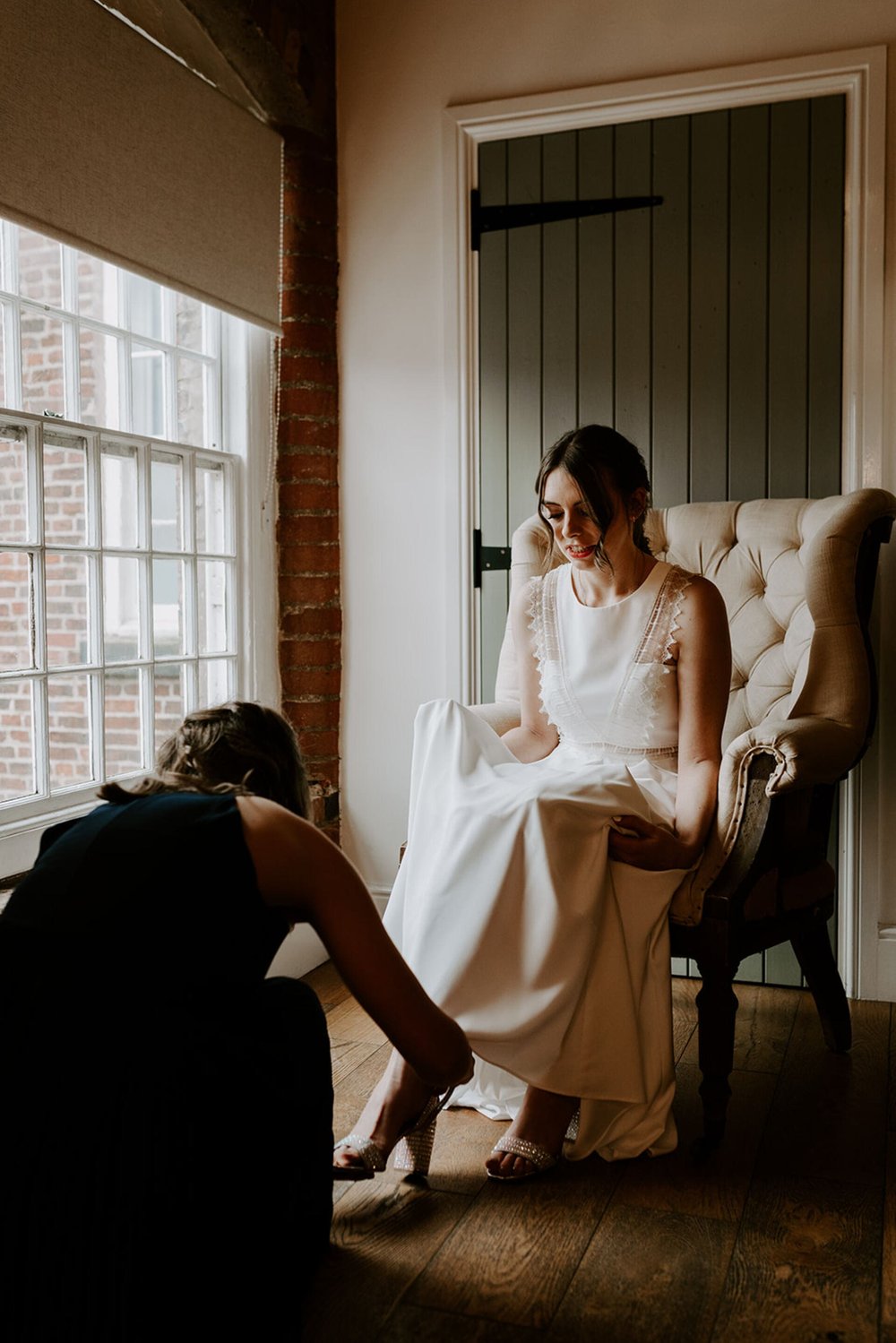  Emma sits in high back winged armchair whilst a bridesmaid does up her shoes. 