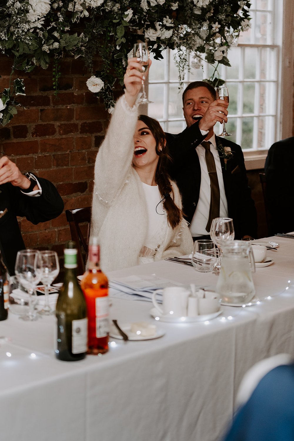  Emma and John sit at their top table. Emma has a loose fitting, ivory wool cardigan over her dress and raises her glass for a toast. 