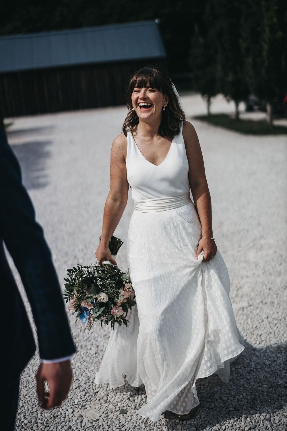  Sarah laughs as she walks outside. Her dress is ivory soft crepe, sleeveless and v neck. She wears a soft chiffon, dotty skirt over her dress with an ivory silk sash tied around her waist. 