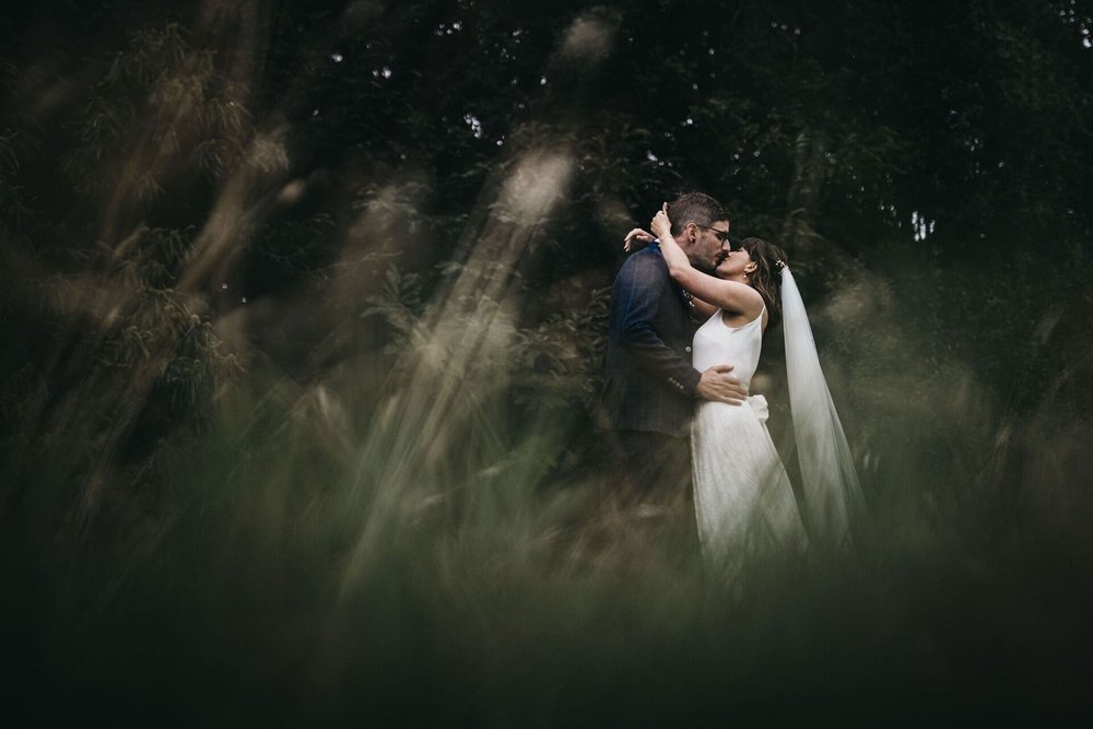  Sarah and Mike share a kiss and embrace. The photo is taken from ground level looking up at them through long grass. 