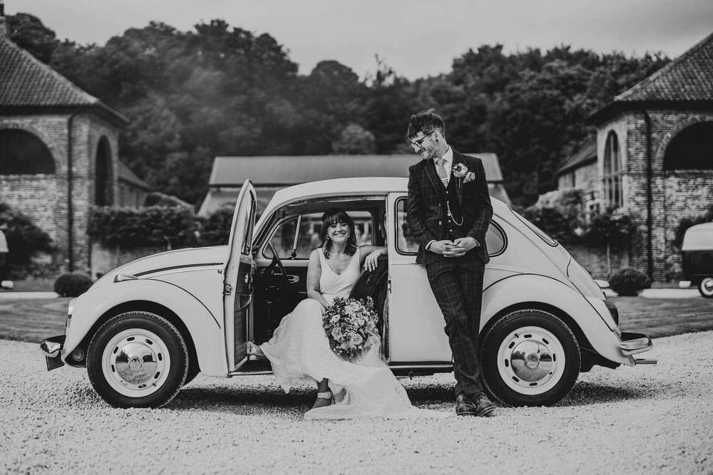  Sarah sits in the passenger seat of an old Beetle car, facing outwards with the door open. Mike leans against the car looking towards her. 