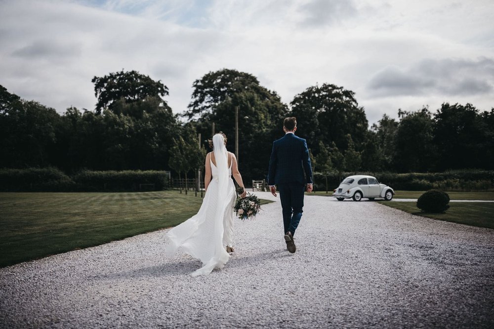  Sarah and Mike walk away from the camera through the grounds of the venue. Sarah wears a train length veil which flows out behind her dress.  