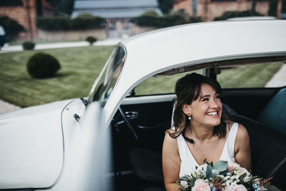  A close up of Sarah sat in the old Beetle car facing outwards with the door open. She’s laughing while holding her bouquet. 