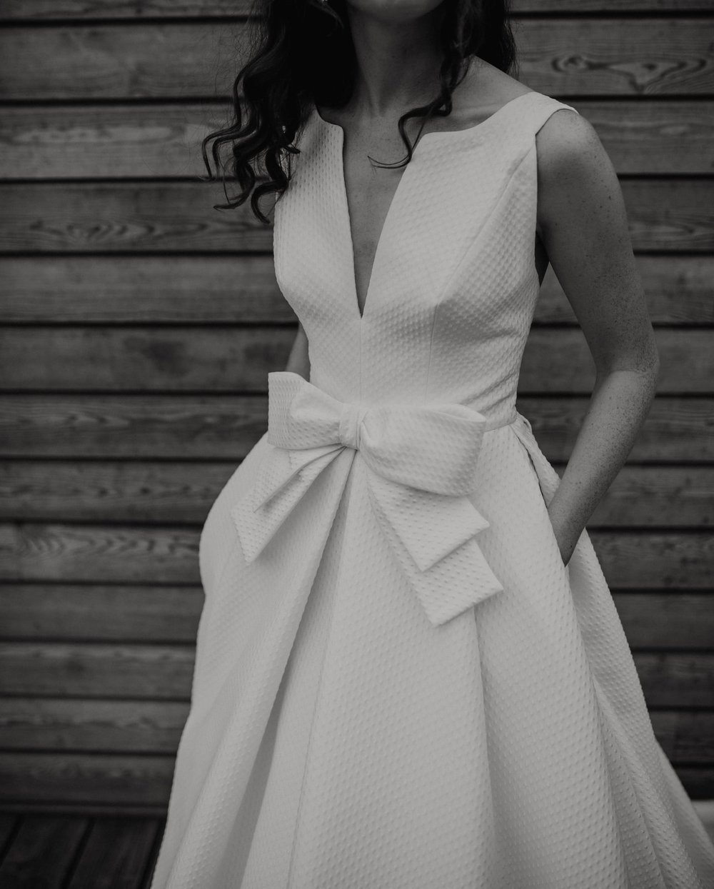  A black and white close up image of Steph in the dress. The dotty texture is clear to see and a large bow adorns the front waist line. The neckline is a deep, narrow V with a soft boat neckline across the shoulders. 