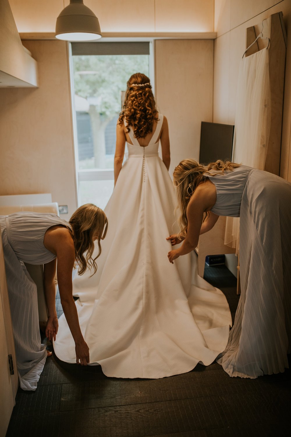  Steph faces away from the camera whist 2 bridesmaids pull out the length of her dress train. The dress is made from a dotty textured taffeta fabric, with a full skirt and large train. There are two wide straps that come down from the shoulders and j