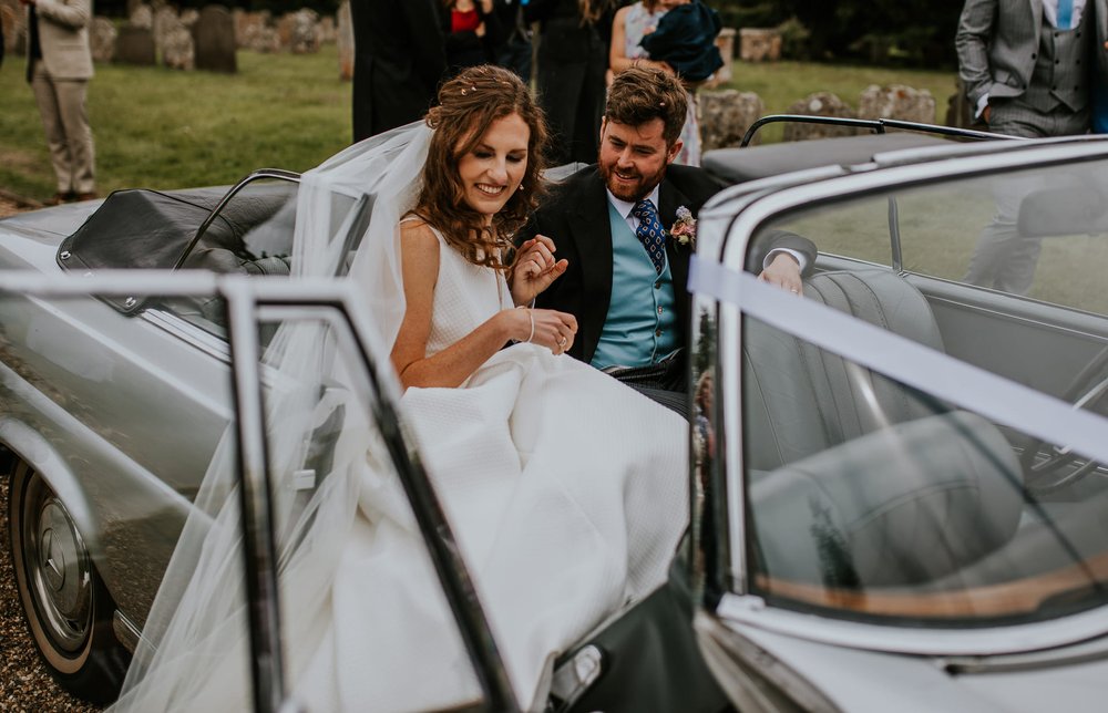  Steph and Tom take a seat in a vintage, soft top silver car. Tom wears a dark suit with blue waistcoat that matches the blue of the bridesmaids. 