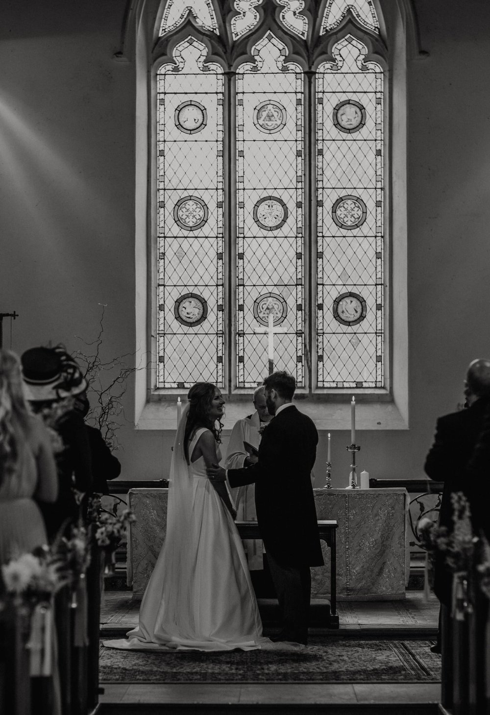  Steph and Tom stand at the alter during the ceremony. Behind them is a large, arched stain glass window made from three panels. 