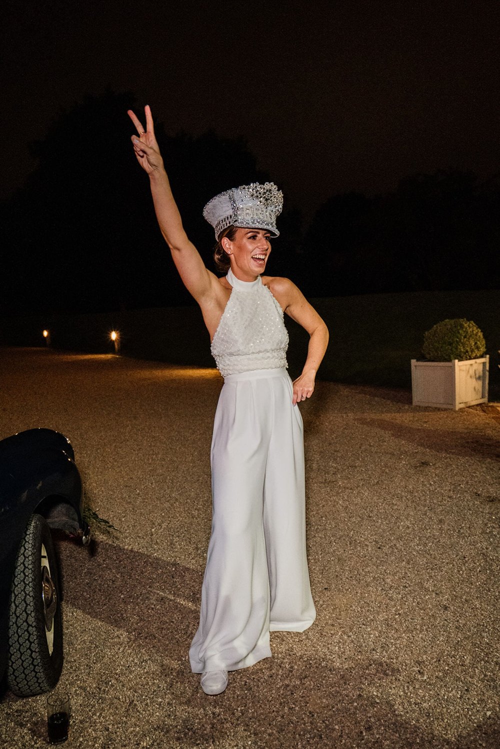  Sophie poses with her arm in the air and other hand on her hip. She wears a bride hat adored with sequins, a halter neck Ivory sequin top and wide leg crepe trousers. It’s evening at the wedding and she is in her second outfit of the day. 