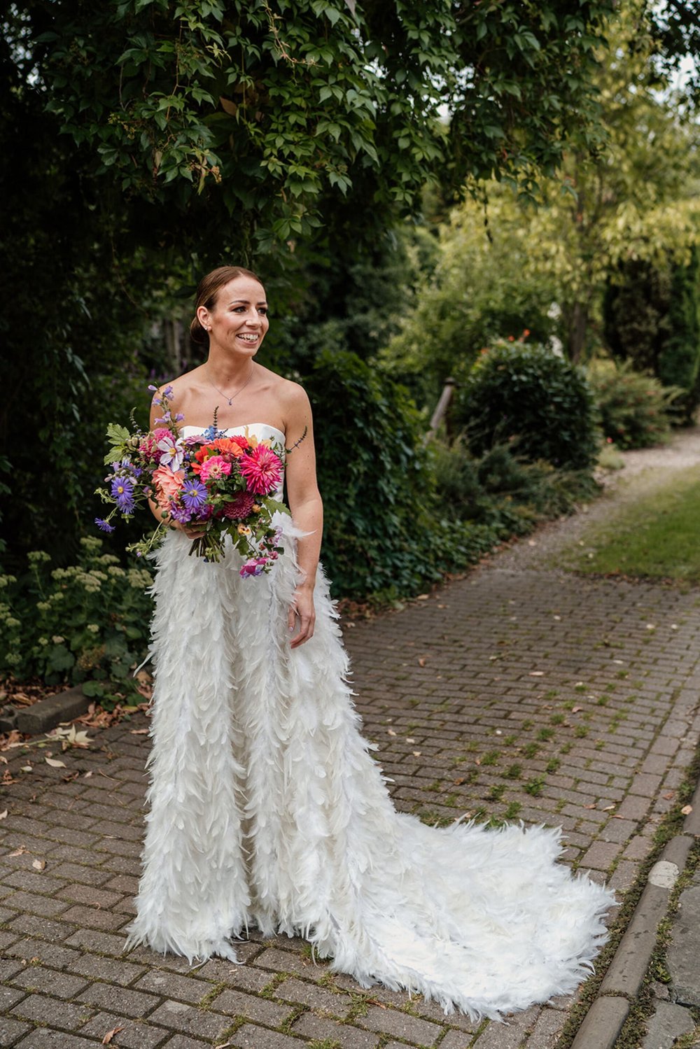  Sophie stands on a cobbled pathway in front of trees and shrubs. She holds her flowers which are bright and bold florals; pinks, purples., corals and green foliage. She wears a strapless satin, structured top that at the waist point joins a full fea