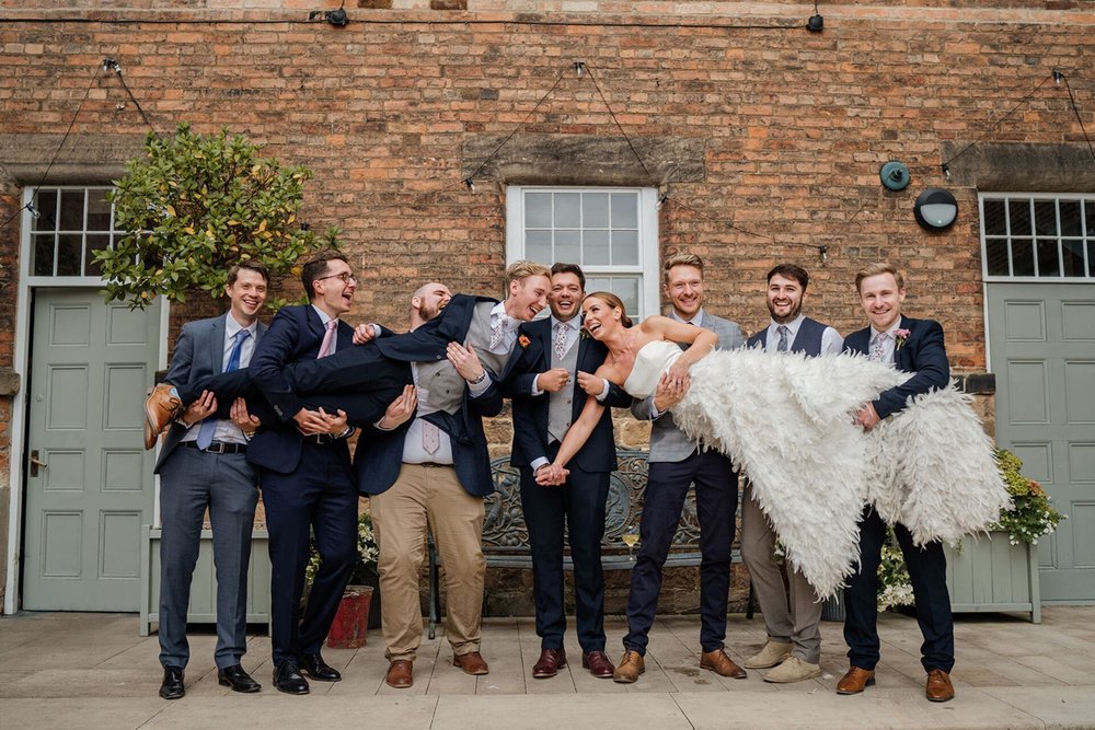  Pete and Sophie are held in the air by 8 guests from the wedding party. They are in front of an old brick building with sage green doors and a decorative bench.  