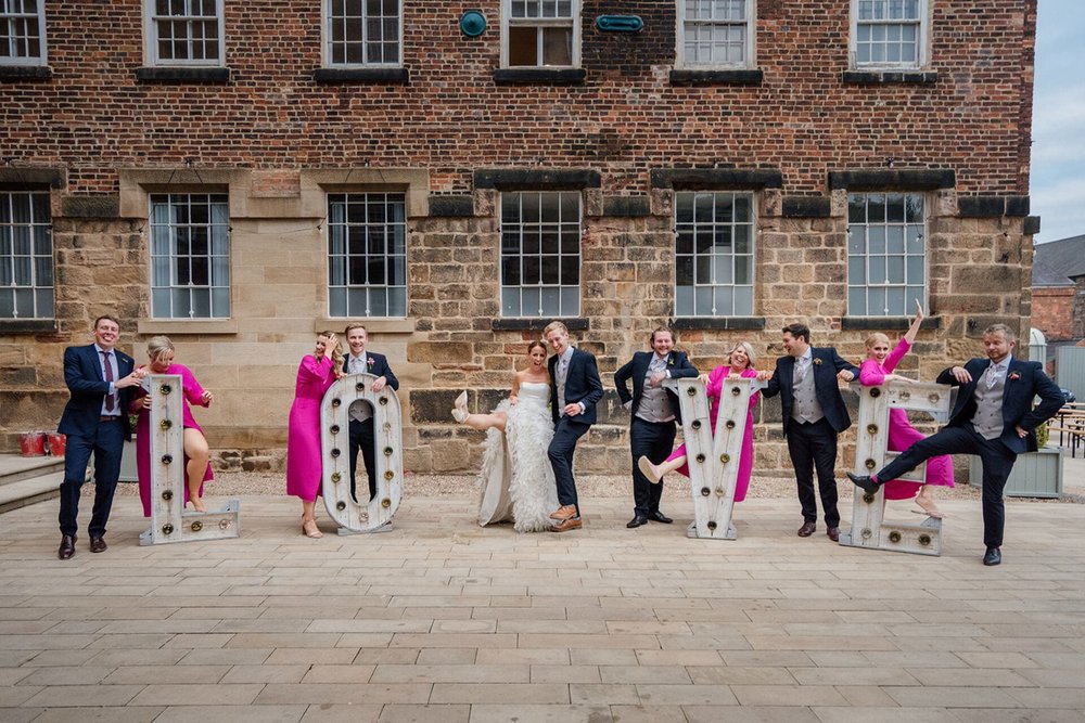  Large light up letters spelling the word "‘LOVE” are outside the venue. Behind each letter stands a bridesmaid in a bright pink dress with one of the ushers. They all pull funny poses and in the middle are Pete and Sophie; Sophie lifts her leg into 
