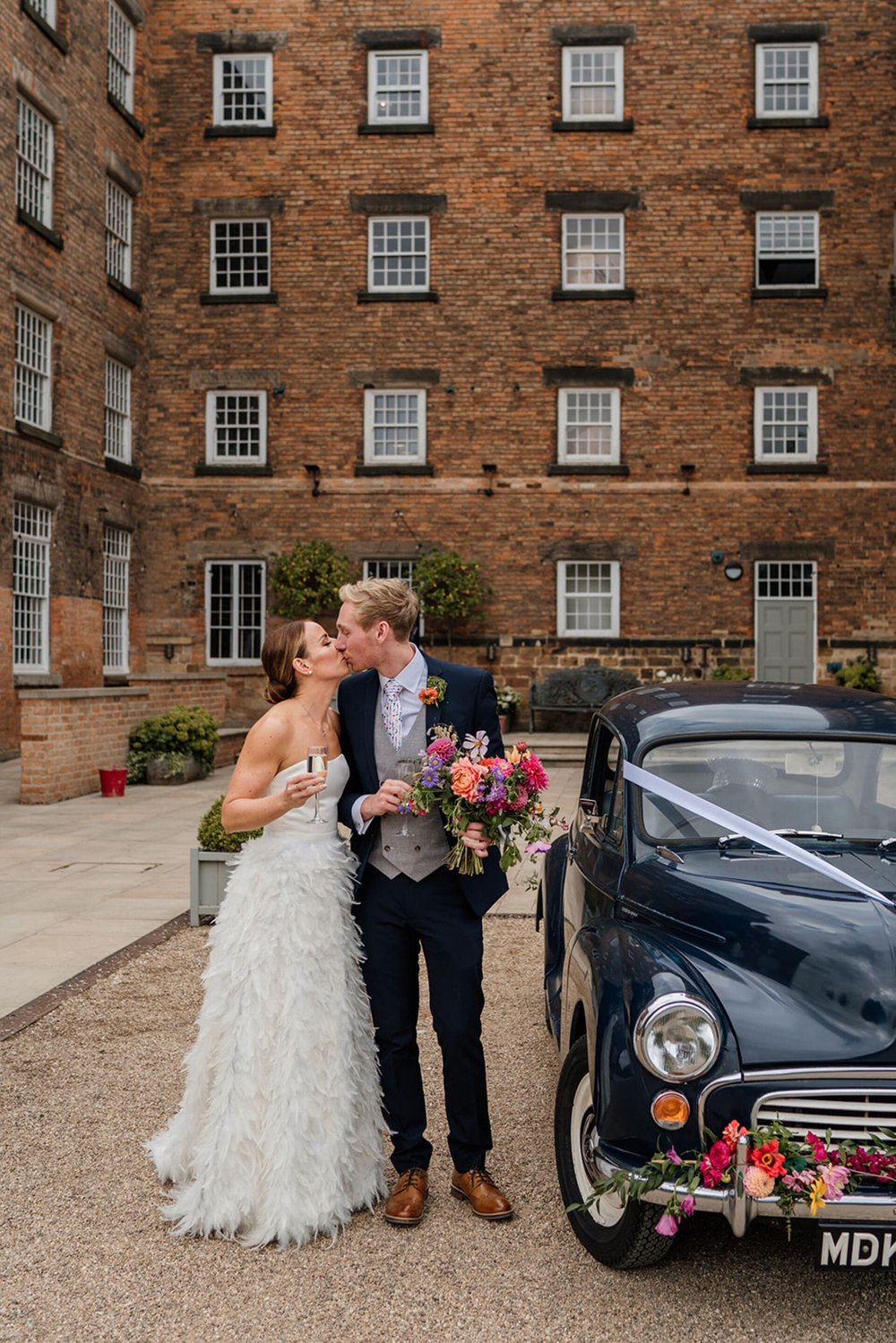  Sophie and Pete share a kiss next to the 1969 Morris Minor navy blue car and in front of the West Mill venue. They both have a glass of bubbly and Pete holds Sophie’s flowers. 