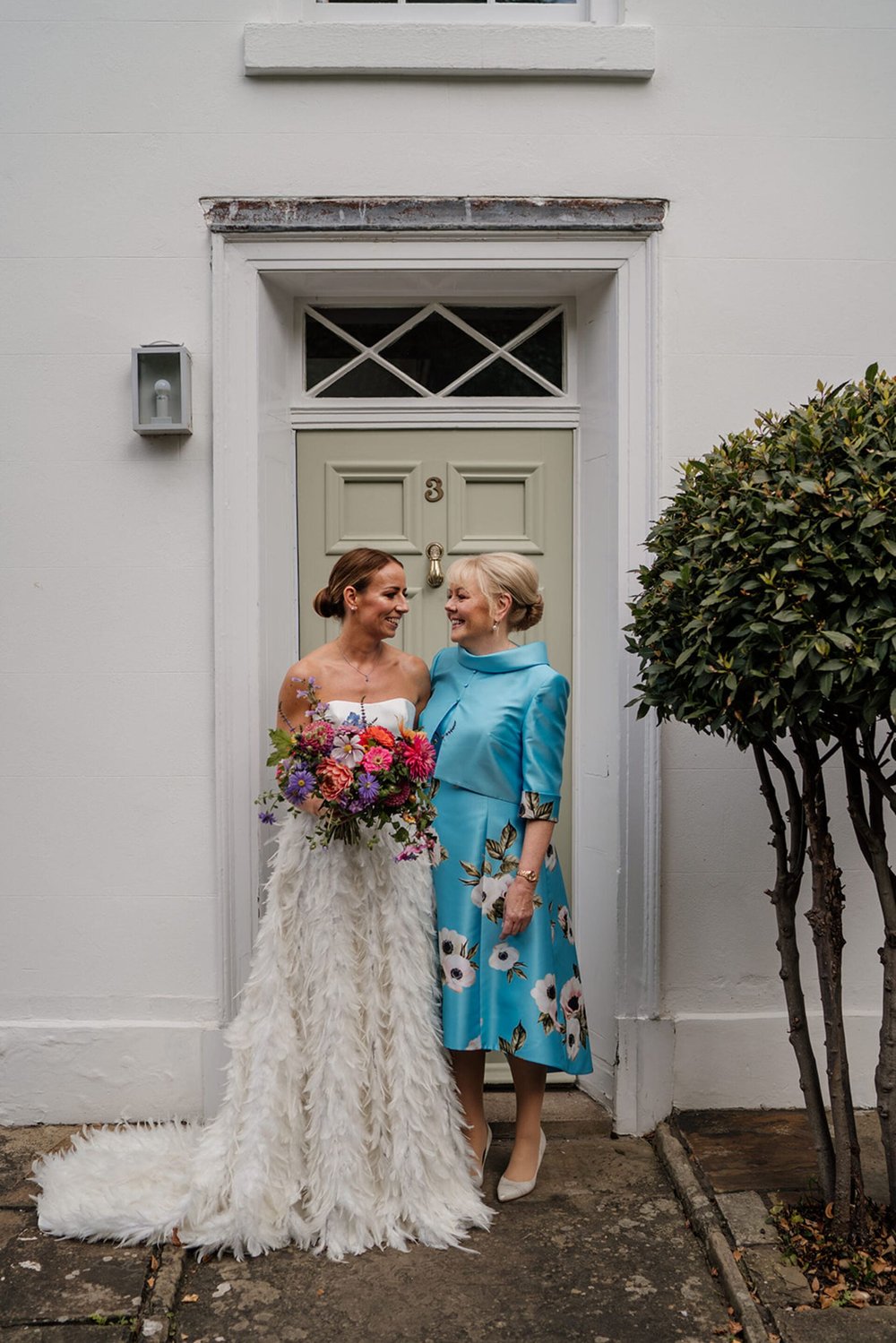  Sophie and her mum stand in front of a doorway of a white Georgian style house. Her mum wears a blue dress with large white flowers, and a high neck satin, 3/4 sleeve blue jacket. 